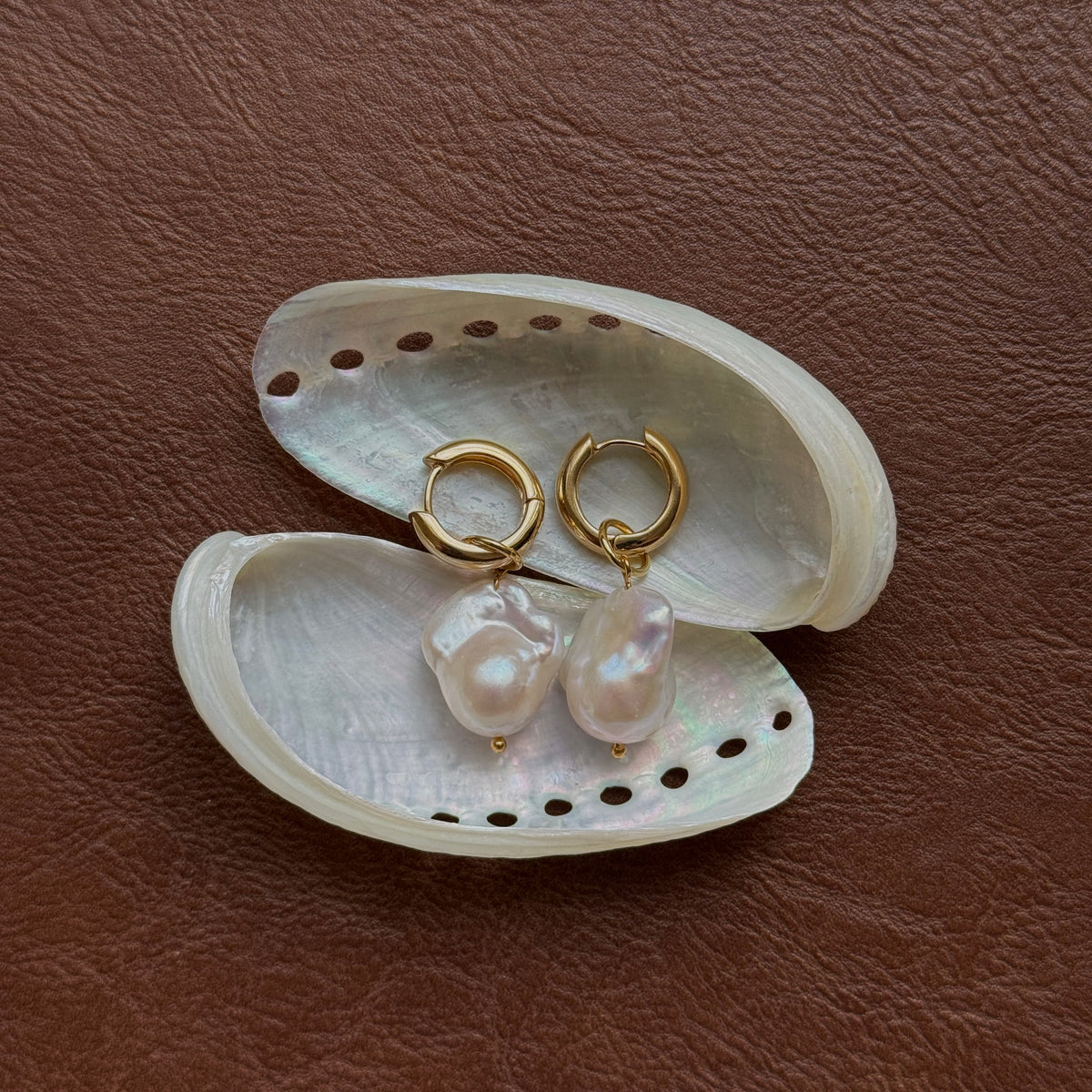 A pair of gold hoop earrings with baroque pearl drops, displayed on a white abalone shell over a brown textured leather background.