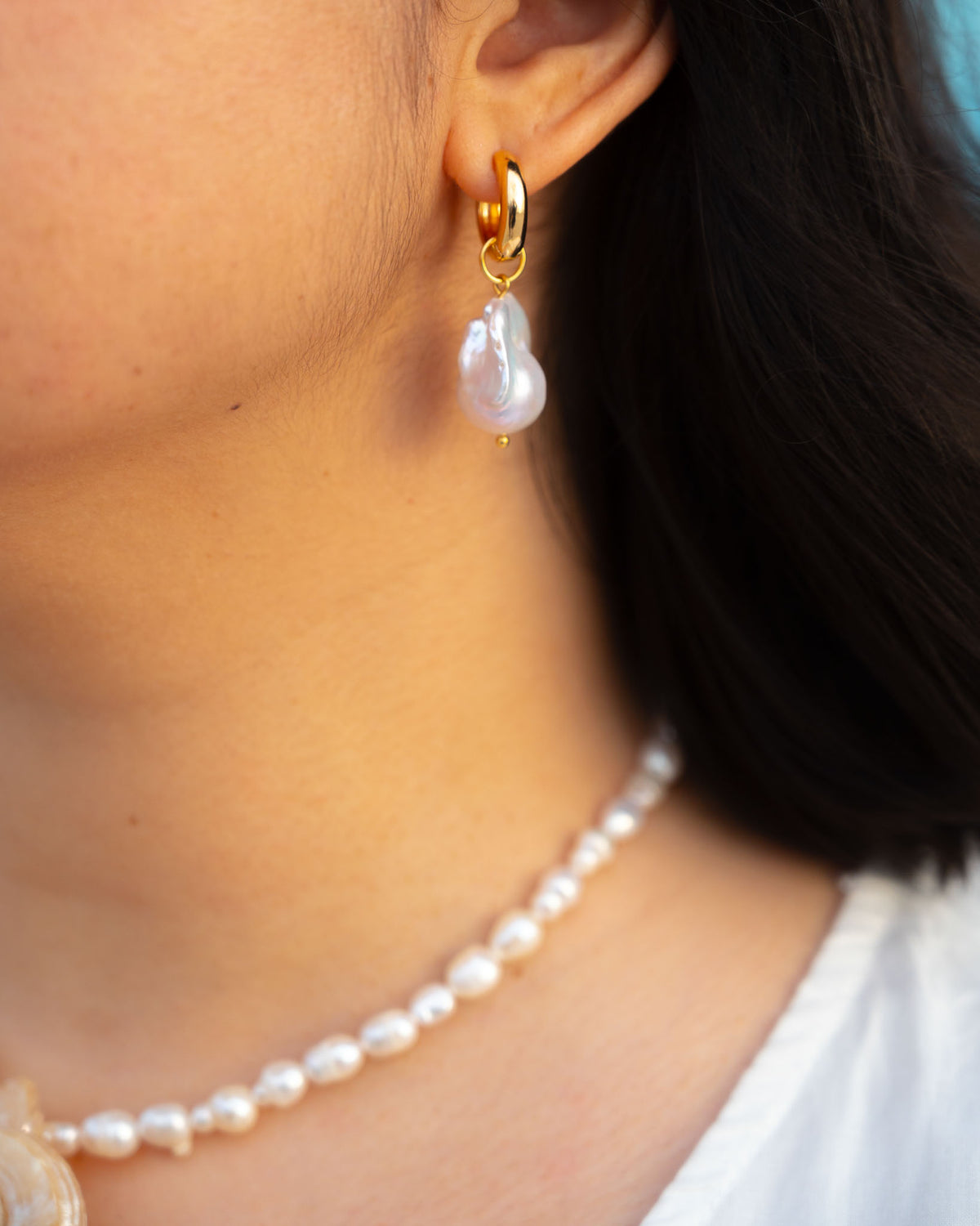 A close-up of a person wearing a gold huggie hoop earring with a dangling baroque pearl, paired with a matching pearl necklace.